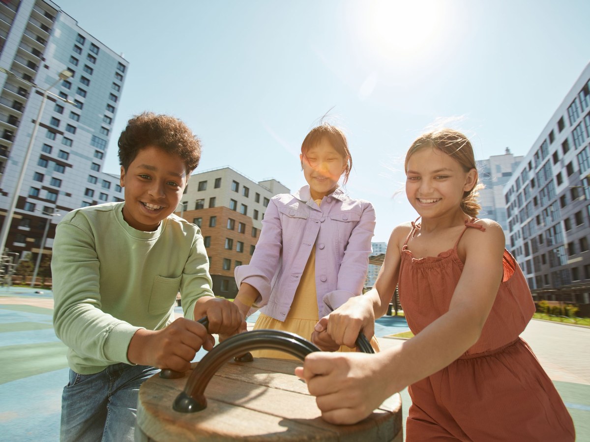 Building social confidence. Three children outdoors working a hand crank together in bright sunlight, leaning in shoulder to shoulder.