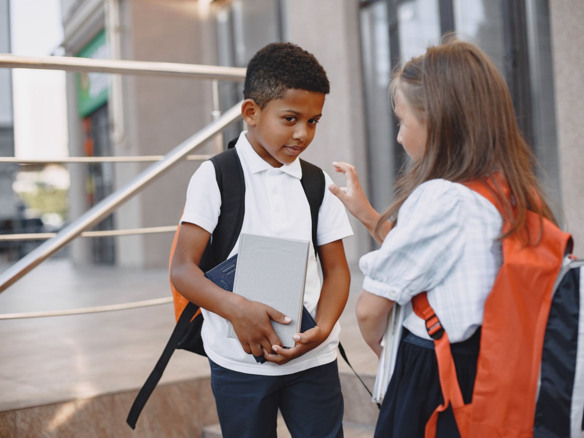 Conversation skills. Two children with backpacks standing face to face, one talking and gesturing while the other listens and holds a notebook.