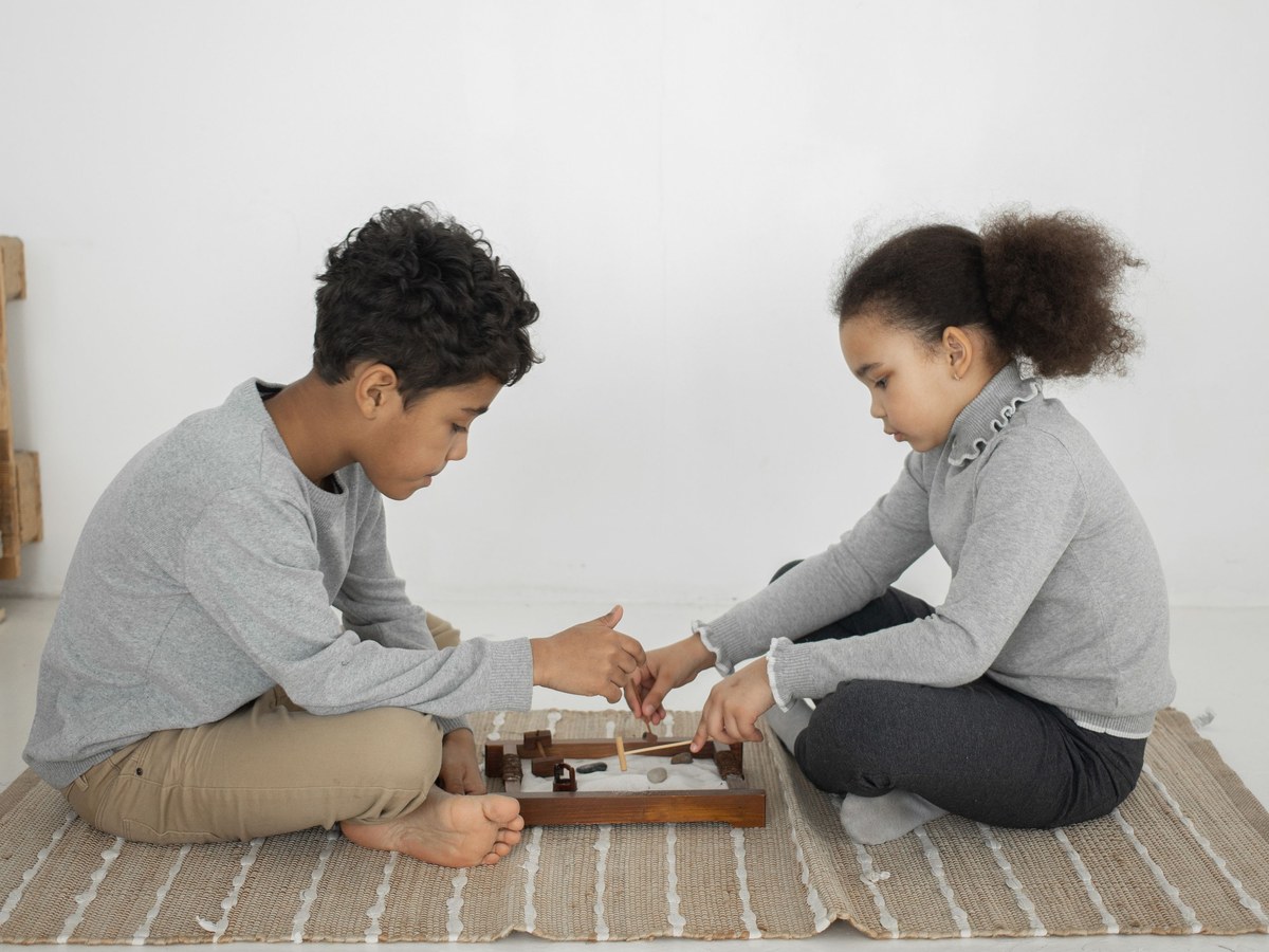 Game play skills. A boy and a girl sitting on a rug facing each other, taking turns at a small wooden board game on the floor between them.