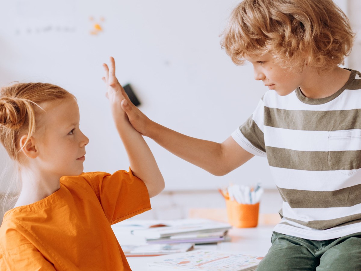 Understanding interpersonal relationships. Two children meeting in a high five across a table in a bright classroom, turned toward each other.