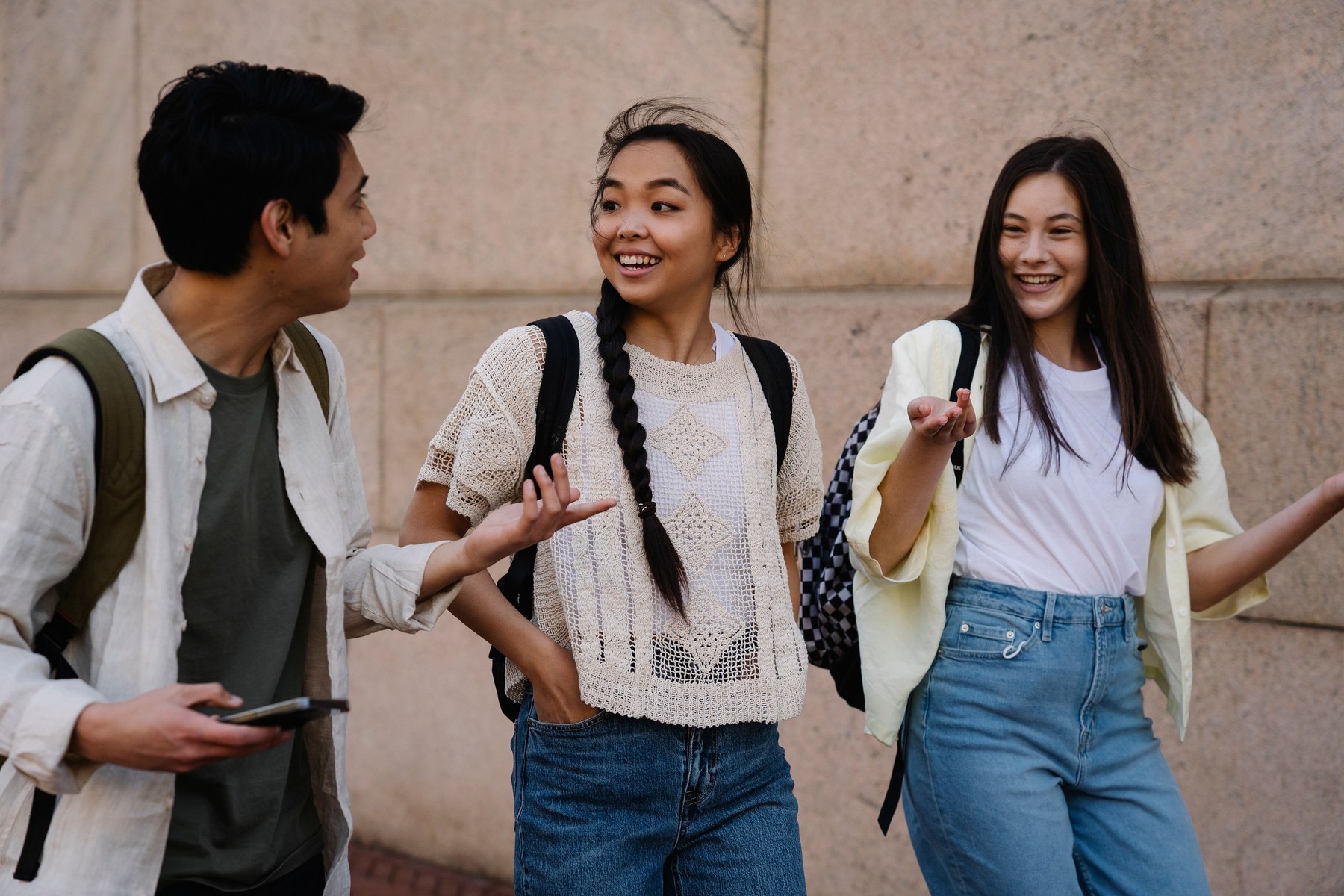 Social skills class for young adults in Great Neck. Three young adults with backpacks standing together outdoors, talking and laughing mid conversation.